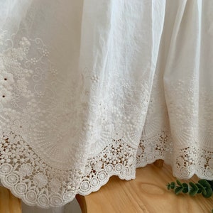 May include: White cotton fabric with delicate embroidered lace trim. The lace features a floral design with scalloped edges.