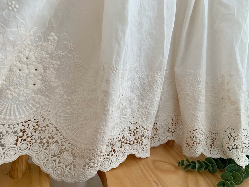 May include: White cotton fabric with delicate embroidered lace trim. The lace features a floral design with scalloped edges.