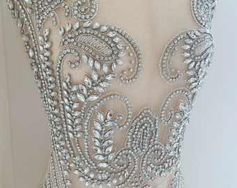 May include: A silver beaded and rhinestone applique with a paisley design. The applique is on a sheer fabric background.