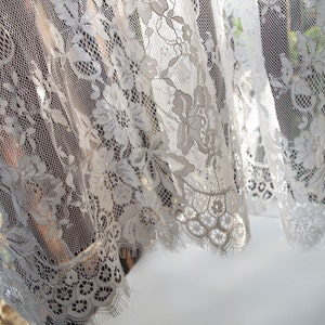 May include: White lace curtain with a floral pattern. The lace is delicate and intricate, with a scalloped edge.