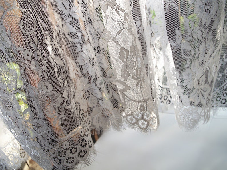 May include: White lace curtain with a floral pattern. The lace is delicate and intricate, with a scalloped edge.