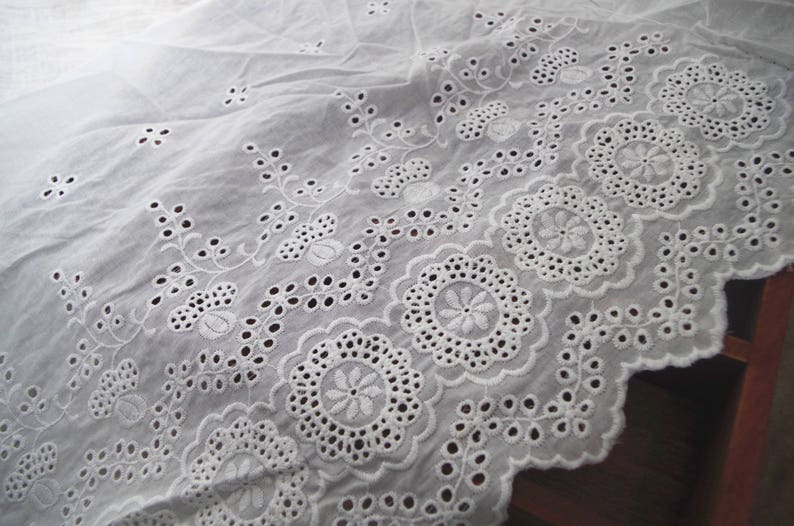 May include: White cotton fabric with a delicate embroidered design featuring floral motifs and scalloped edges.