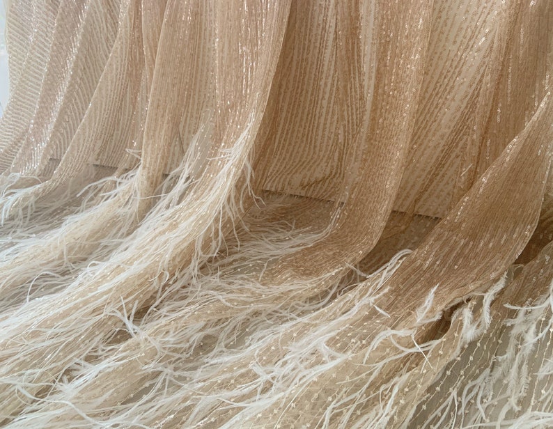 K&ouml;nnte beinhalten: Beigefarbener Stoff mit glitzernder Textur und wei&szlig;em Federnbesatz. Der Stoff ist durchsichtig und hat eine zarte, flie&szlig;ende Optik.