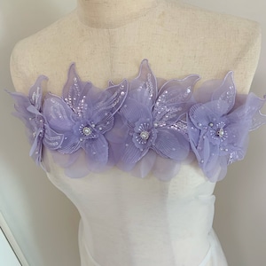 Può includere: Applicazione floreale lavanda con abbellimenti di paillettes e perline. L'applicazione presenta cinque grandi disegni floreali a strati con petali delicati e un gioiello centrale. I fiori sono attaccati a un tessuto bianco trasparente.