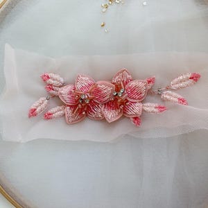 May include: A decorative floral appliqué featuring two central pink and white beaded flowers with clear crystal centres, surrounded by smaller beaded leaf-shaped accents in matching pink and white tones, all mounted on a piece of sheer white fabric.