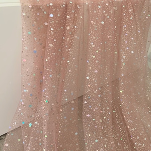 May include: A sheer, blush pink fabric with scattered, iridescent sequins. The fabric is draped, creating soft folds and shadows. The sequins catch the light, creating a sparkling effect. This material is suitable for dresses, veils, or decorative projects.
