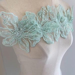 Könnte beinhalten: Eine hellgrüne florale Spitzenapplikation mit Perlenbesatz. Die Applikation ist aus zartem Stoff gefertigt und zeigt eine Ansammlung von Blumen.