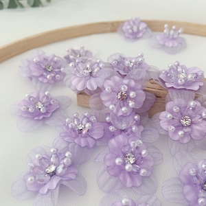 Könnte beinhalten: Eine Sammlung von lavendelfarbenen Stoffblumen mit Perlen- und Strassbesatz. Die Blumen sind auf einer Holzoberfläche und einem weißen Hintergrund angeordnet.