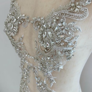 May include: A white lace appliqué featuring intricate silver beadwork and clear faceted crystals arranged in floral and vine patterns. The decorative piece is designed with a sheer mesh base and is displayed on a neutral-toned mannequin torso.