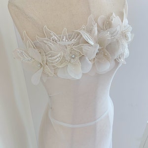 Puede incluir: Un vestido blanco transparente con un delicado adorno floral en el corpiño. Las flores están hechas de tela blanca y tienen un brillo sutil.