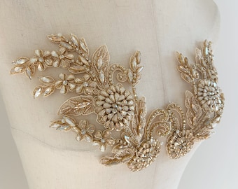 May include: Gold beaded applique with a floral design. The applique features a large central flower with smaller flowers and leaves surrounding it. The applique is perfect for adding a touch of sparkle and elegance to any garment.