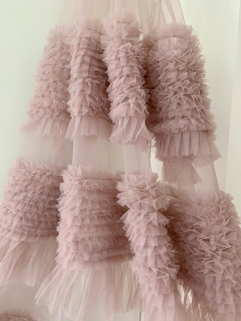 May include: A dusty rose tulle fabric with multiple layers of ruffles. The fabric is soft and delicate, and the ruffles are evenly spaced.