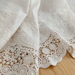 May include: White cotton fabric with delicate embroidery and a lace trim. The fabric is folded over, showing the intricate details of the embroidery and lace.