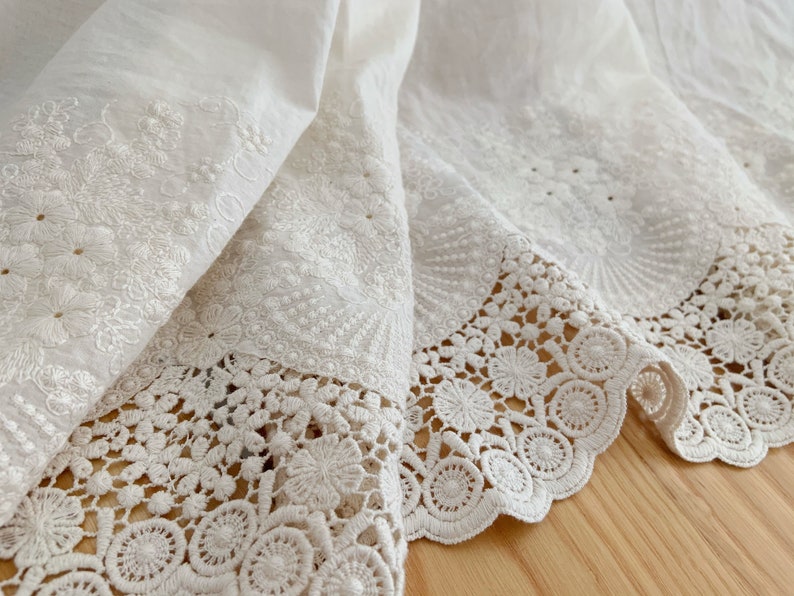 May include: White cotton fabric with delicate embroidery and a lace trim. The fabric is folded over, showing the intricate details of the embroidery and lace.