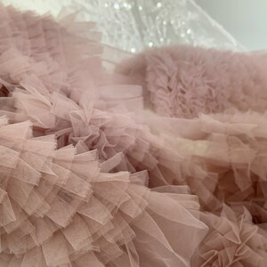 May include: Close-up of a blush pink tulle fabric with multiple layers of ruffles. The fabric is soft and delicate, with a slightly textured surface.