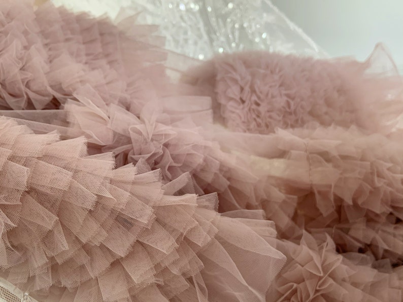 May include: Close-up of a blush pink tulle fabric with multiple layers of ruffles. The fabric is soft and delicate, with a slightly textured surface.