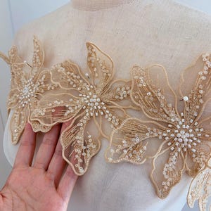 May include: A beige floral lace trim with pearl embellishments. The lace is delicate and intricate, with a floral pattern and small pearls scattered throughout. The trim is perfect for adding a touch of elegance to any project.