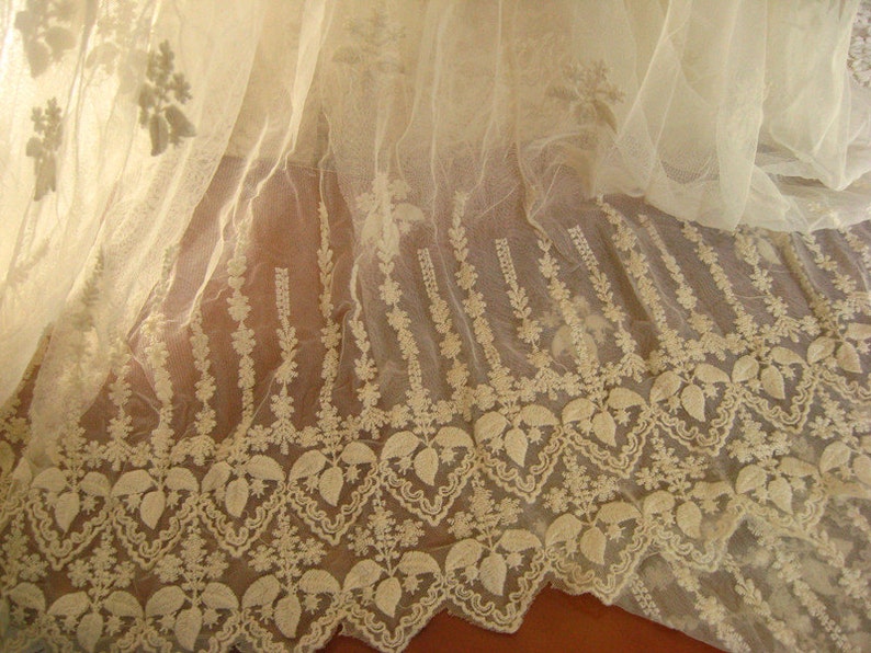 May include: White lace fabric with a delicate floral pattern. The lace has a scalloped edge and is sheer and lightweight.