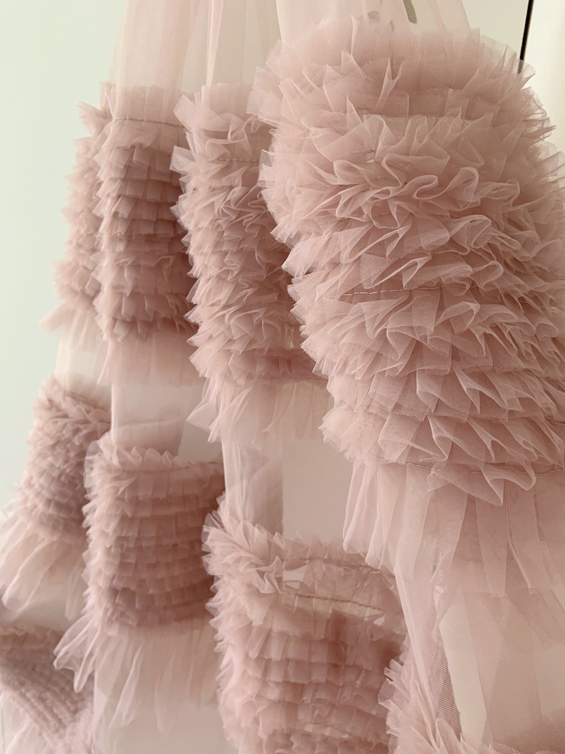May include: A blush pink tulle fabric with multiple layers of ruffles. The fabric is soft and delicate, and the ruffles are arranged in a cascading pattern.