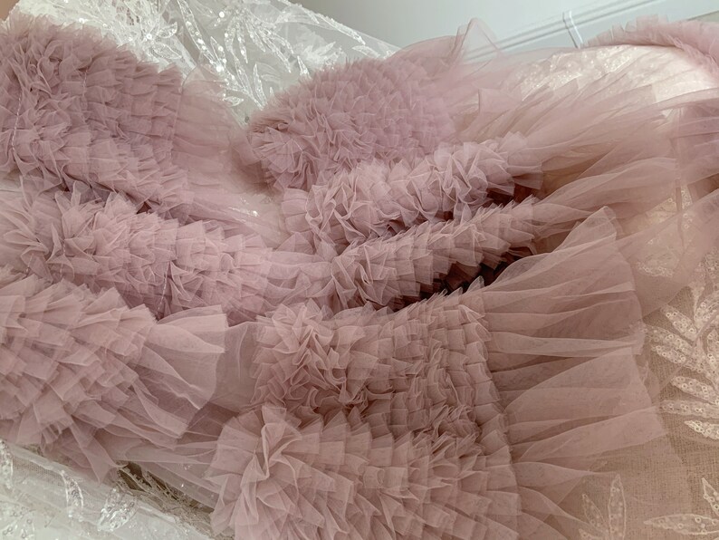 May include: A close-up of a dusty rose tulle fabric with multiple layers of ruffles. The fabric is soft and delicate, with a flowing texture.