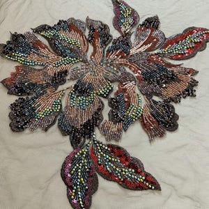 May include: An intricate floral applique featuring dense embroidery with iridescent beads and sequins in shades of copper, bronze, dark gray, and deep red. The design is composed of multiple petal and leaf shapes arranged on a sheer beige mesh fabric.