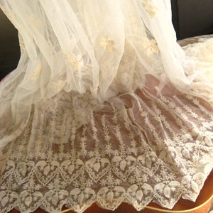 May include: White lace fabric with a delicate floral pattern. The lace is draped over a brown wooden surface.