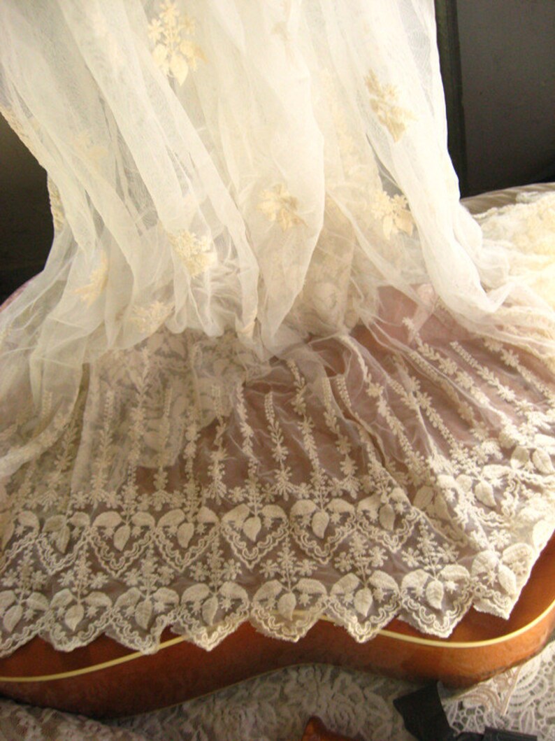 May include: White lace fabric with a delicate floral pattern. The lace is draped over a brown wooden surface.