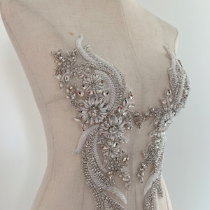 Könnte beinhalten: Dekorative Applikation mit floralem Design, verziert mit glitzernden Strasssteinen, klaren Kristallen und weißen Perlen. Das symmetrische Design ist an einer Schaufensterpuppe befestigt.