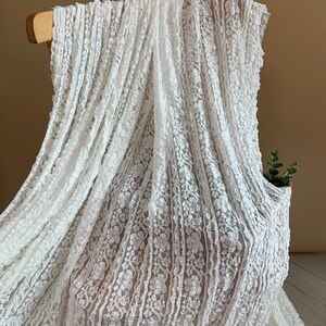 May include: White lace fabric draped over a wooden chair. The delicate fabric features a floral pattern and vertical stripes, creating a textured appearance. The lace is a bright white colour, and the background is a neutral brown.