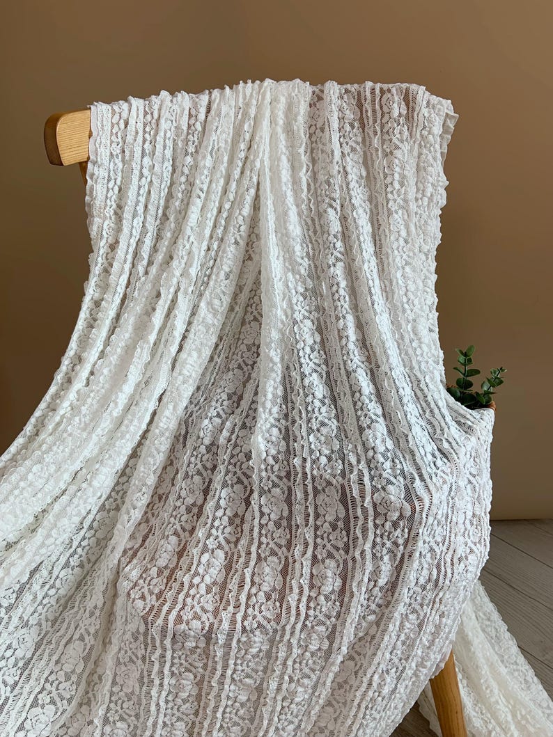 May include: White lace fabric draped over a wooden chair. The delicate fabric features a floral pattern and vertical stripes, creating a textured appearance. The lace is a bright white colour, and the background is a neutral brown.