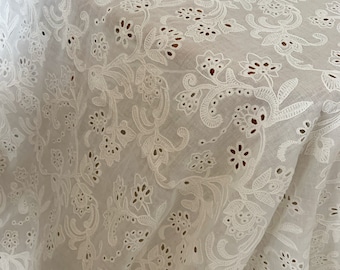 May include: White cotton fabric with a delicate floral eyelet embroidery pattern. The fabric is lightweight and sheer, making it perfect for summer garments.