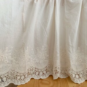 May include: White cotton fabric with a delicate embroidered lace trim. The lace features a scalloped edge and intricate floral designs.