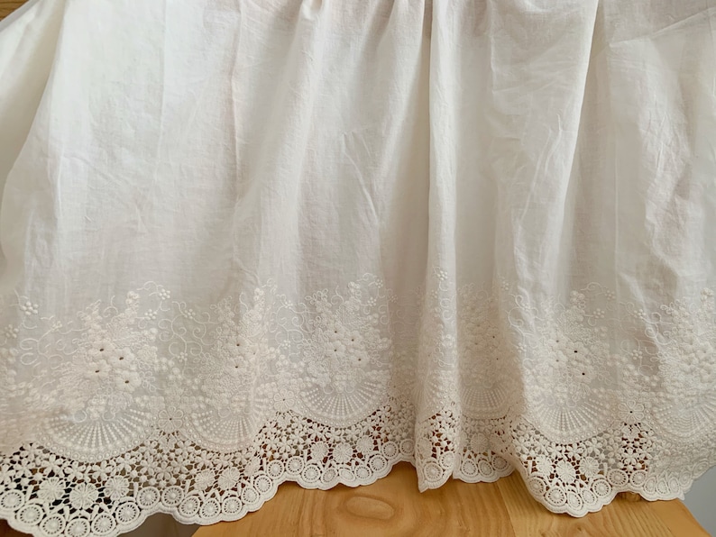 May include: White cotton fabric with a delicate embroidered lace trim. The lace features a scalloped edge and intricate floral designs.
