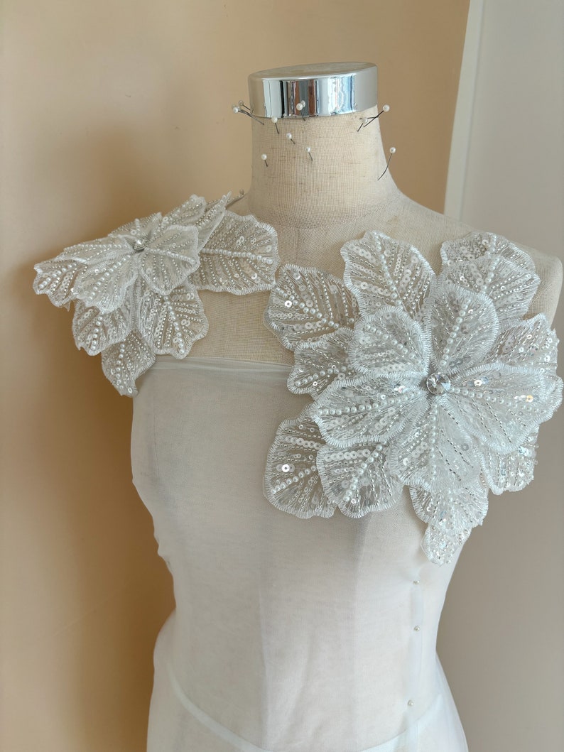 Pu&ograve; includere: Due applicazioni floreali bianche con decorazioni di perle e paillettes. Le applicazioni sono su uno sfondo di tessuto bianco.