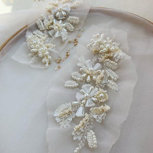Könnte beinhalten: Zwei elfenbeinfarbene florale Applikationen aus transparentem Tüll, verziert mit weißen Saatperlen, glänzenden Perlen und plissierten Bandakzenten. Die komplexen Designs zeigen geschichtete Blütenblätter und perlenbesetzte Stiele.