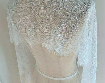 May include: White lace fabric with a delicate floral pattern. The lace has a scalloped edge and is shown draped over a dress form.