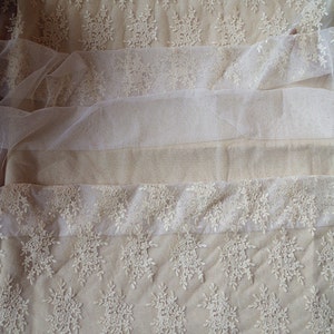 May include: Close-up of cream-colored lace fabric with floral and snowflake patterns. The fabric is layered with different textures, including a sheer tulle. The bottom edge features a scalloped lace trim. Ideal for bridal or craft projects.