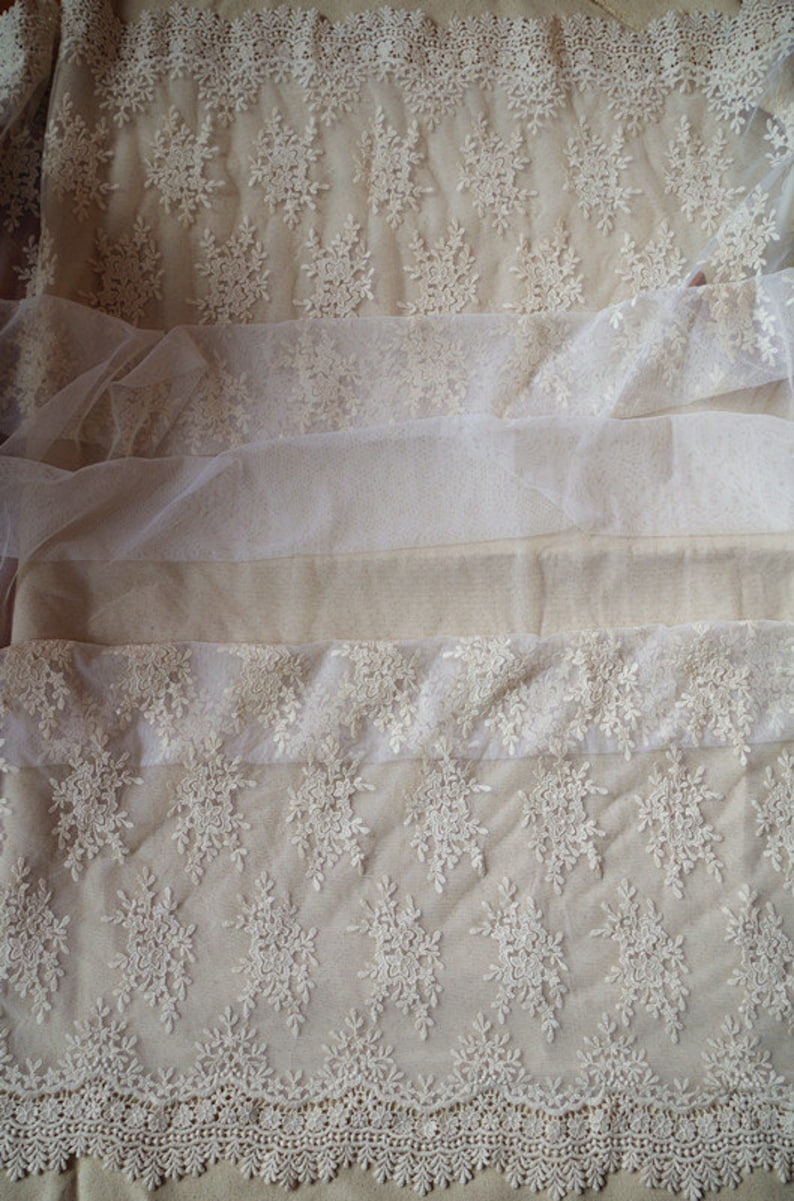 May include: Close-up of cream-colored lace fabric with floral and snowflake patterns. The fabric is layered with different textures, including a sheer tulle. The bottom edge features a scalloped lace trim. Ideal for bridal or craft projects.