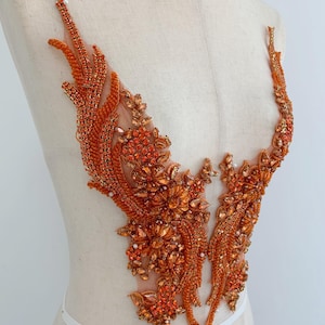 Può includere: Applicazione di perline arancioni con un design floreale e a forma di piuma. L'applicazione è realizzata con perline e strass arancioni di varie dimensioni, cuciti su un supporto in rete trasparente. Il design è simmetrico e sembra essere per l'abbellimento.