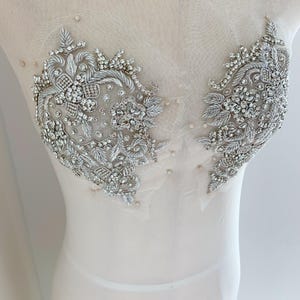 以下が含まれることがあります： 銀とクリスタルで刺繍された対称的なアップリケが2つ付いたブライダルボディス。アップリケは、複雑なビーズ細工と輝くクリスタルを備えた花と葉のデザインが特徴です。ボディスは、透け感のあるオフホワイトの生地で作られています。
