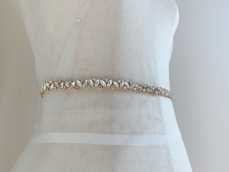 Puede incluir: Un cintur&oacute;n de novia dorado con adornos de cristal transparente. El cintur&oacute;n est&aacute; hecho de un material delgado y flexible y est&aacute; adornado con una fila de cristales brillantes.
