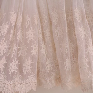 May include: Off-white lace fabric with a floral pattern. The delicate lace features a repeating design of stylized flowers and leaves, creating a textured and elegant appearance. The fabric is semi-sheer, allowing light to pass through.