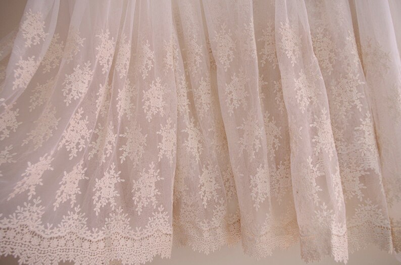 May include: Off-white lace fabric with a floral pattern. The delicate lace features a repeating design of stylized flowers and leaves, creating a textured and elegant appearance. The fabric is semi-sheer, allowing light to pass through.