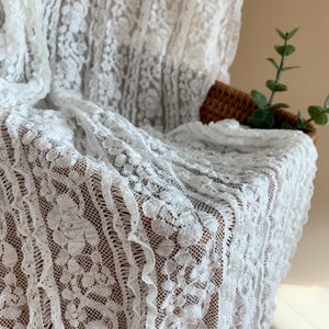 May include: White lace fabric with a floral pattern. The delicate material is draped over a surface, showcasing the intricate details of the design. The lace has a scalloped edge and a textured appearance.