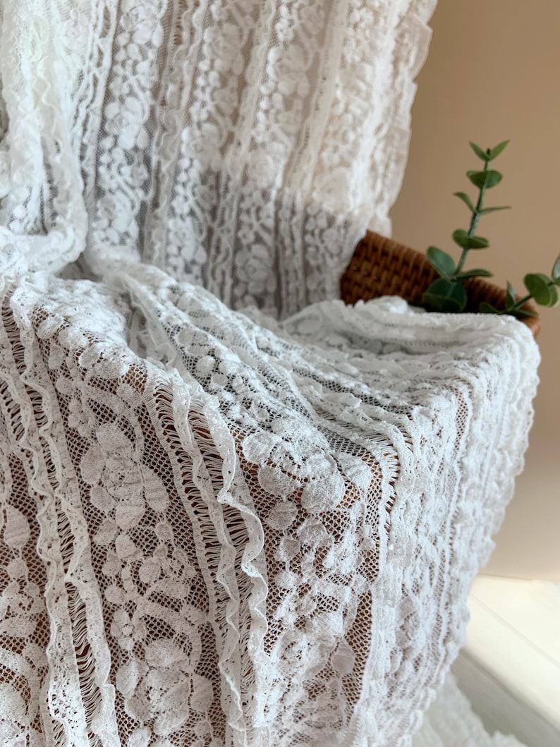 May include: White lace fabric with a floral pattern. The delicate material is draped over a surface, showcasing the intricate details of the design. The lace has a scalloped edge and a textured appearance.
