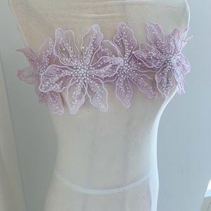May include: A delicate, floral applique featuring five lavender-colored flowers. Each flower is intricately detailed with white sequins and beads, creating a shimmering effect. The applique is designed to be sewn onto fabric, adding a touch of elegance to garments or accessories.