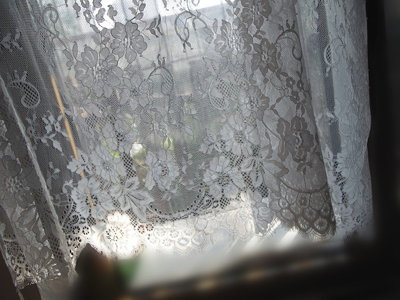 May include: White lace curtain with a floral pattern, hanging in a window. The lace is delicate and intricate, and the light shines through it, creating a soft, ethereal glow.