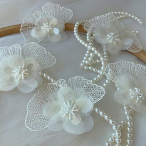 Op de afbeelding: Close-up van ivoorkleurige bloemversieringen met delicate, gelaagde bloemblaadjes en parelaccenten. De bloemen zijn verbonden door strengen kleine, witte parels. Het ontwerp bevat een kantachtig materiaal rond de bloemblaadjes.