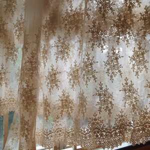 May include: Sheer, beige lace curtain with a floral snowflake pattern. The curtain has a scalloped edge and hangs in front of a window, allowing light to filter through. The intricate design adds a touch of elegance to any room.