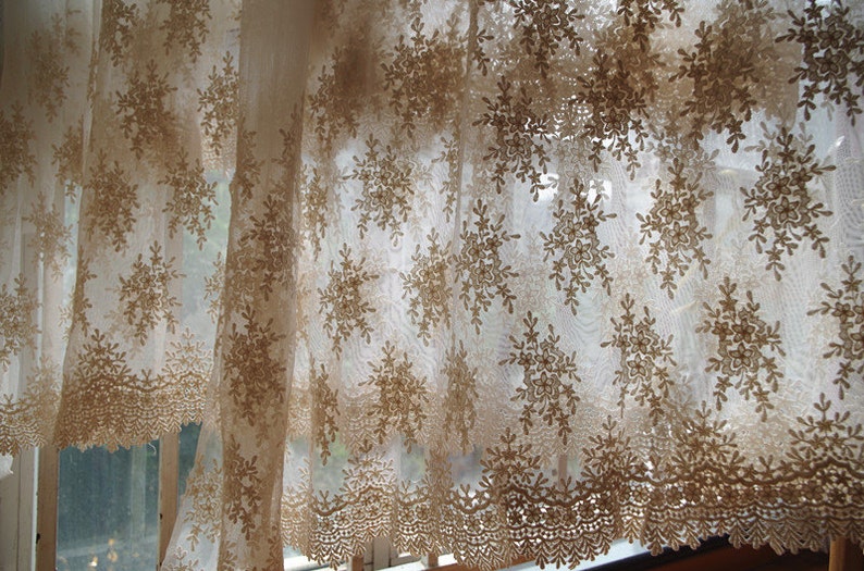 May include: Sheer, beige lace curtain with a floral snowflake pattern. The curtain has a scalloped edge and hangs in front of a window, allowing light to filter through. The intricate design adds a touch of elegance to any room.